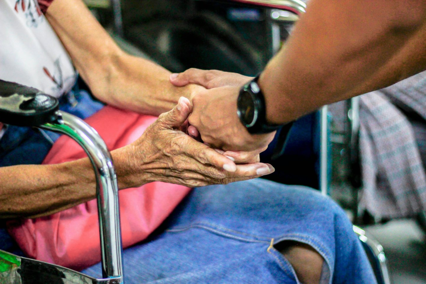 Young hands hold elderly wrinkled hands in a wheelchair.