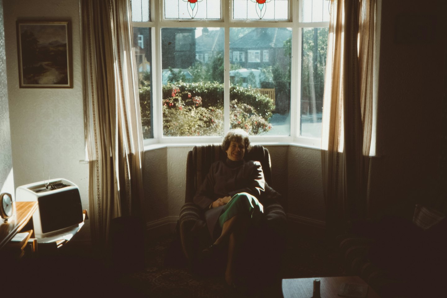 An elderly woman sits smiling in an armchair by a window.